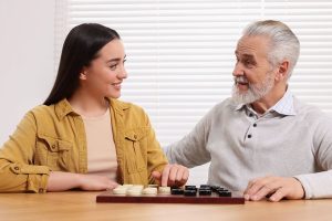 Playing checkers. Senior man learning young woman at table in room